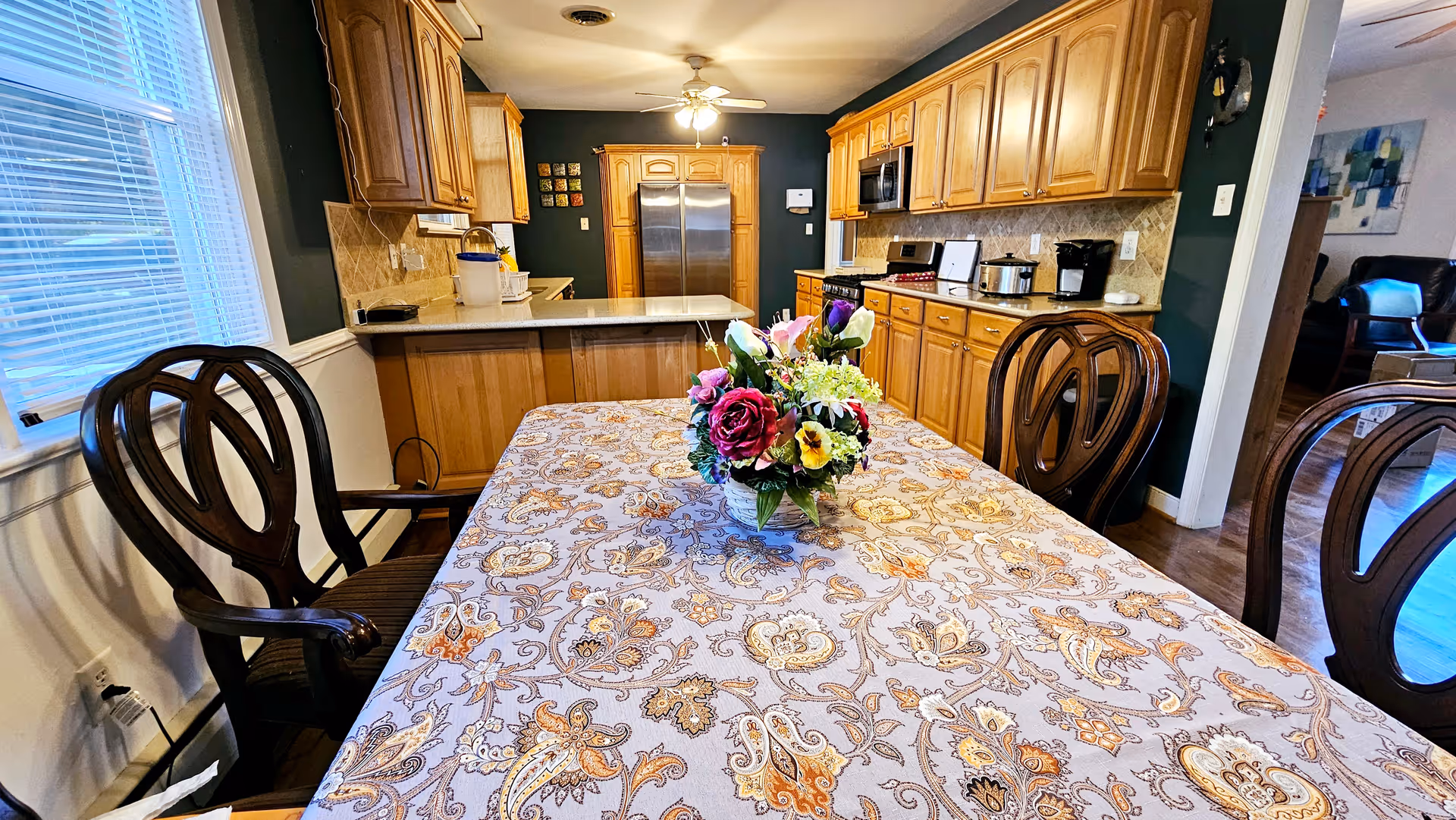 A kitchen and dining area with wooden cabinets and stainless steel appliances. A table covered with a patterned tablecloth and a floral centerpiece is in the foreground, surrounded by wooden chairs. The kitchen has a refrigerator, microwave, stove, and coffee maker. There is a window with blinds on the left side and a ceiling fan with lights above.
