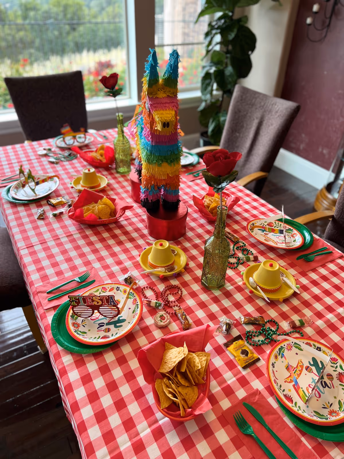 A dining table set for a festive celebration with a red and white checkered tablecloth, colorful plates with fiesta-themed designs, green plastic cutlery, yellow sombrero-shaped cups, bowls of tortilla chips, a colorful llama piñata centerpiece, red rose decorations in green glass bottles, and festive beads and candy scattered on the table. The setting is in a room with large windows and upholstered chairs around the table.