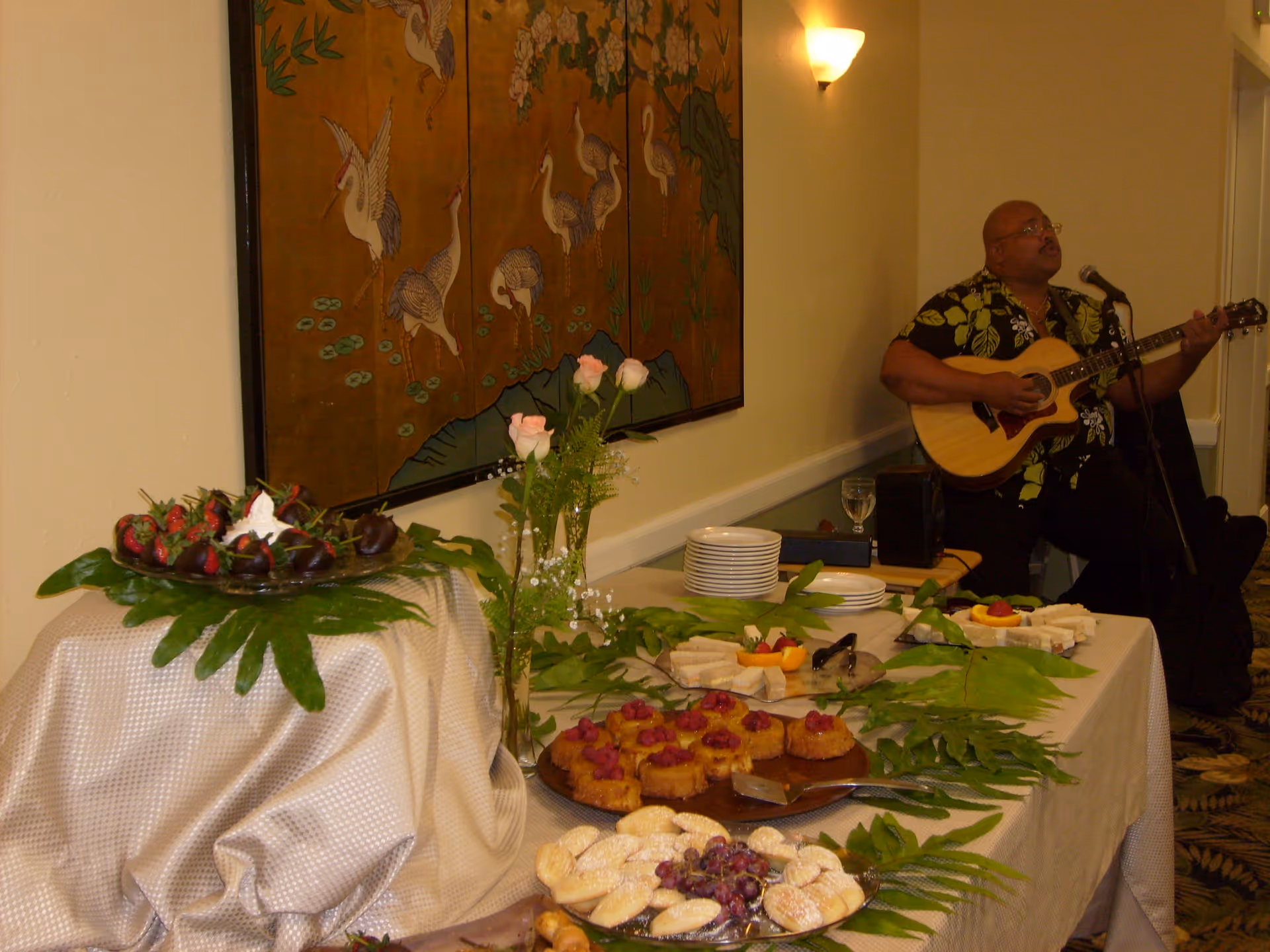 Buffet table with assorted desserts, plates and floral decorations in a room while a musician plays guitar in the background.