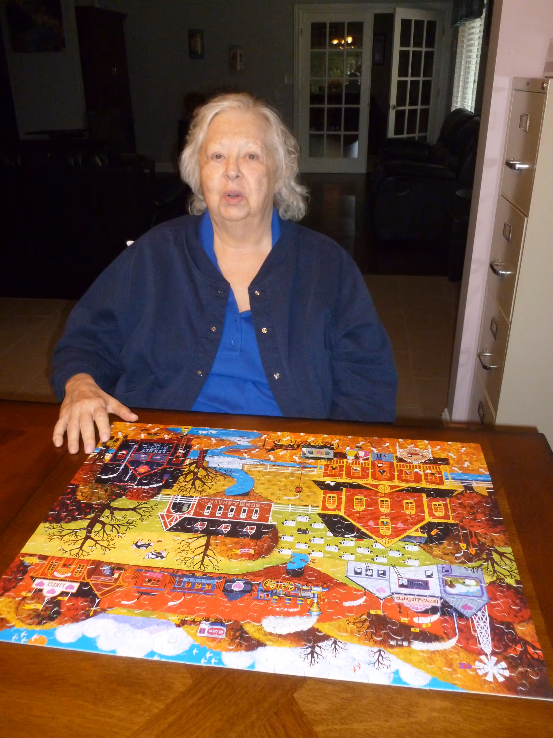 An elderly woman with white hair wearing a blue shirt and dark jacket sits at a wooden table with a colorful completed jigsaw puzzle depicting a farm scene with buildings, trees, and animals. The background shows a dimly lit room with a glass-paneled door and some furniture.