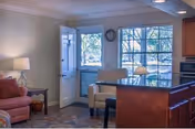 Interior view of a dining hall at Florida Living Retirement Community featuring a cozy seating area with a red armchair and a beige armchair near a window and an open door. The room has a tiled floor, a table with a lamp, and kitchen cabinetry visible on the right side.