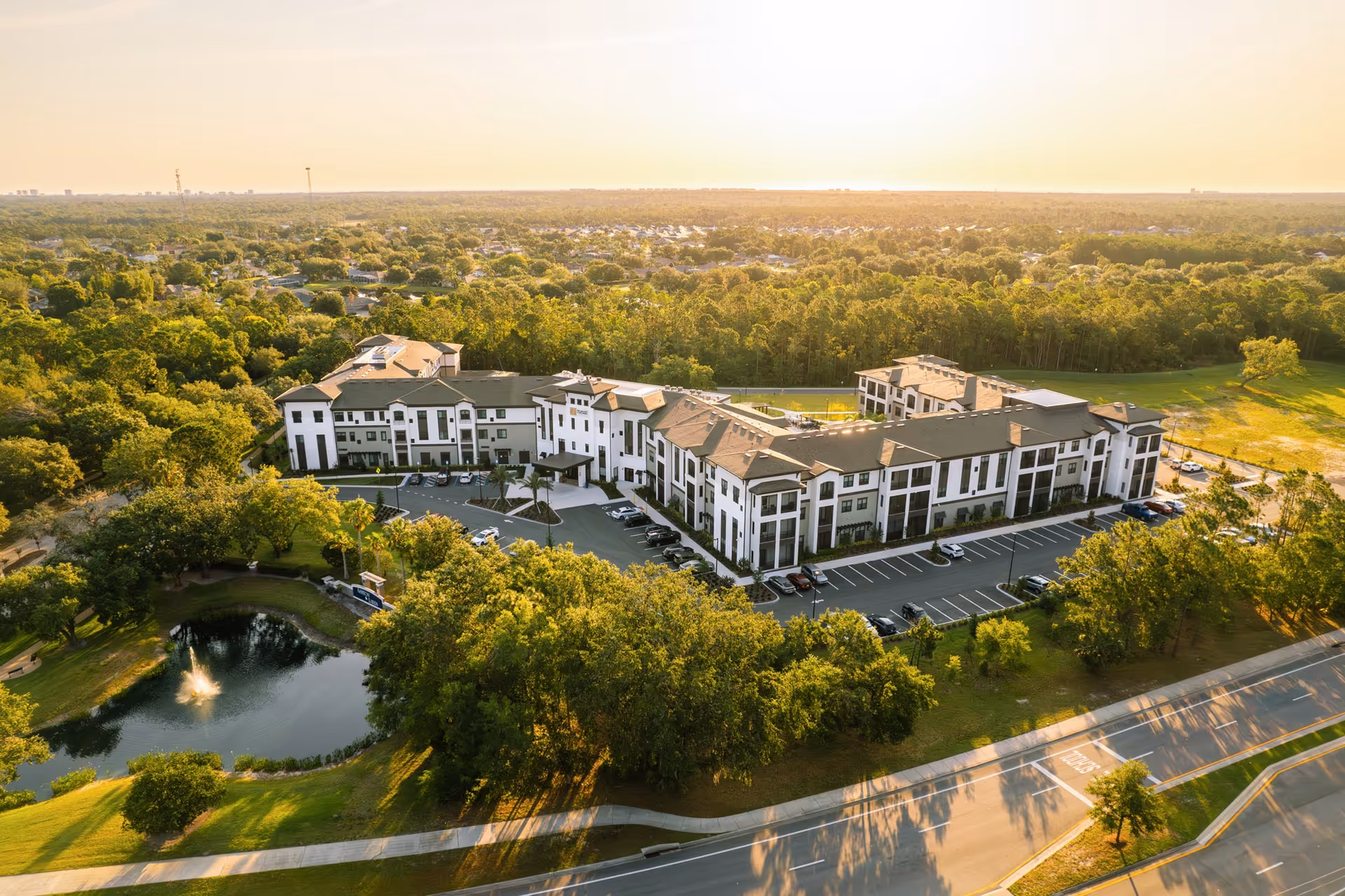 Aerial view of a large, modern senior living facility named Experience Senior Living surrounded by trees and greenery. The building is white with dark accents and has multiple floors with a parking lot in front. There is a small pond with a fountain and a walking path nearby. The scene is bathed in warm sunlight, suggesting early morning or late afternoon.