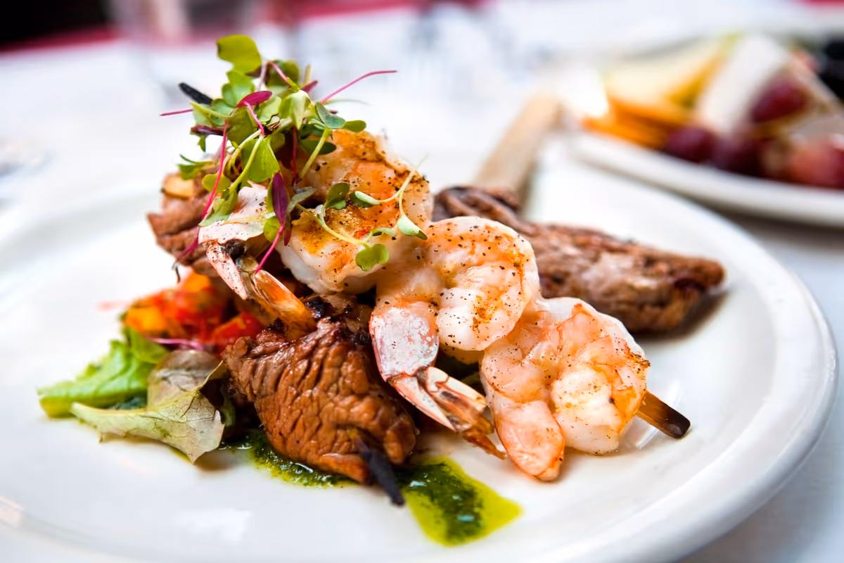 A close-up of a gourmet dish featuring grilled shrimp and steak skewers garnished with fresh microgreens, served on a white plate with a side of leafy greens and a drizzle of green sauce.