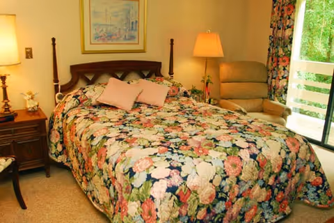 A bedroom with a floral patterned bedspread on a double bed, two pink pillows, a wooden headboard, a nightstand with a lamp and decorative items, an upholstered armchair, floral curtains, and a sliding glass door leading outside.