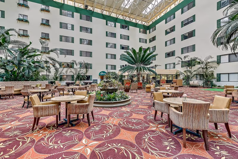 Spacious indoor common area with patterned carpet, multiple round tables with chairs, large potted plants, and a high ceiling with skylights. The area is surrounded by several floors of windows and balconies.