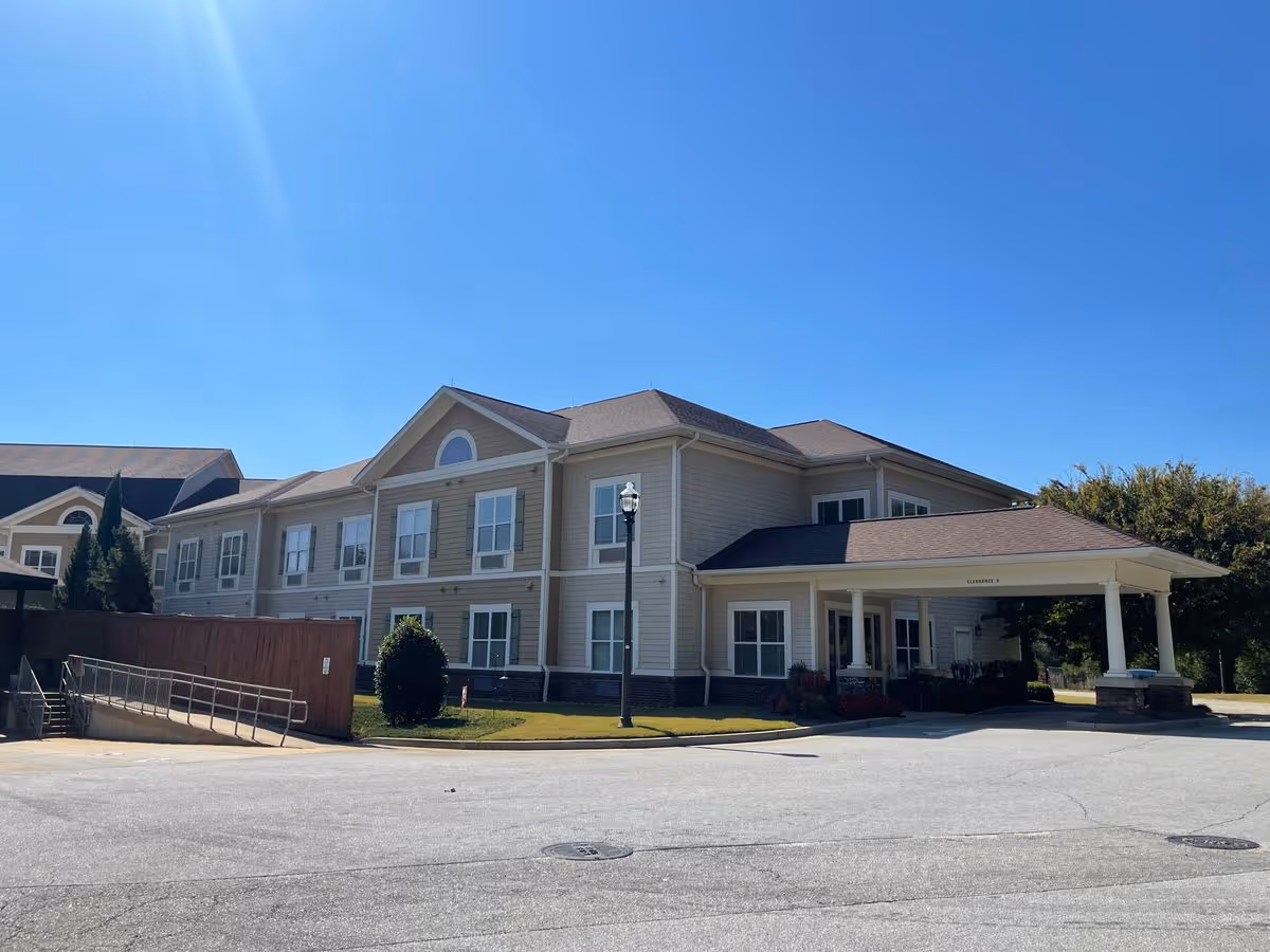 Two-story beige senior living building with a covered entrance/porte-cochère and surrounding driveway under a clear blue sky.