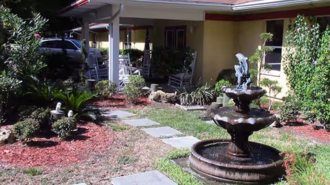 Outdoor garden area at Noble House of Jacksonville featuring a stone pathway leading to a covered porch with white rocking chairs. The garden includes a tiered water fountain, various plants, shrubs, and mulch beds.