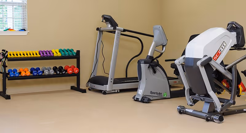 A fitness room with a rack of colorful dumbbells, a treadmill, a stationary bike, and a SciFit exercise machine against beige walls and a window with white blinds.