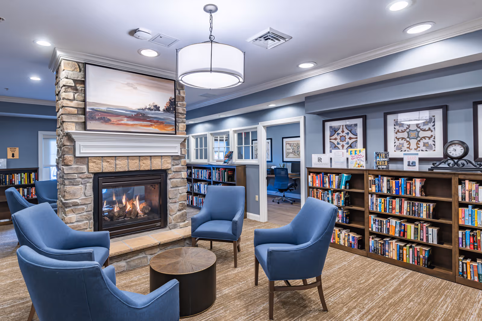 A cozy senior living common room with a stone fireplace, four blue armchairs around a round coffee table, and bookshelves along the wall.
