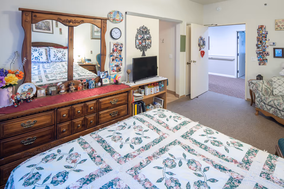 A cozy bedroom in an assisted living facility featuring a bed with a floral quilt, a wooden dresser with a large mirror, various decorative items, a small TV on a stand with shelves holding bottles and books, and an open door leading to a hallway. The room also has a floral patterned armchair and several photo collages on the walls.