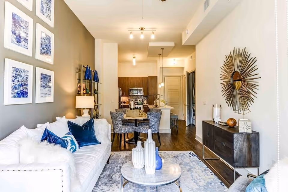 Open-plan living room and kitchen featuring a white sofa with blue pillows, a round coffee table, dining area, and modern wall decor.