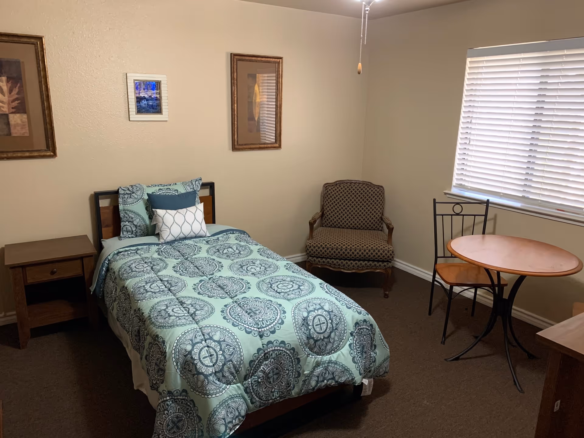 A small bedroom with a single bed covered in a blue and white patterned comforter, a wooden nightstand with a drawer, a cushioned armchair with a patterned fabric, a round wooden table with two chairs, and a window with closed blinds. Three framed pictures hang on the beige walls.