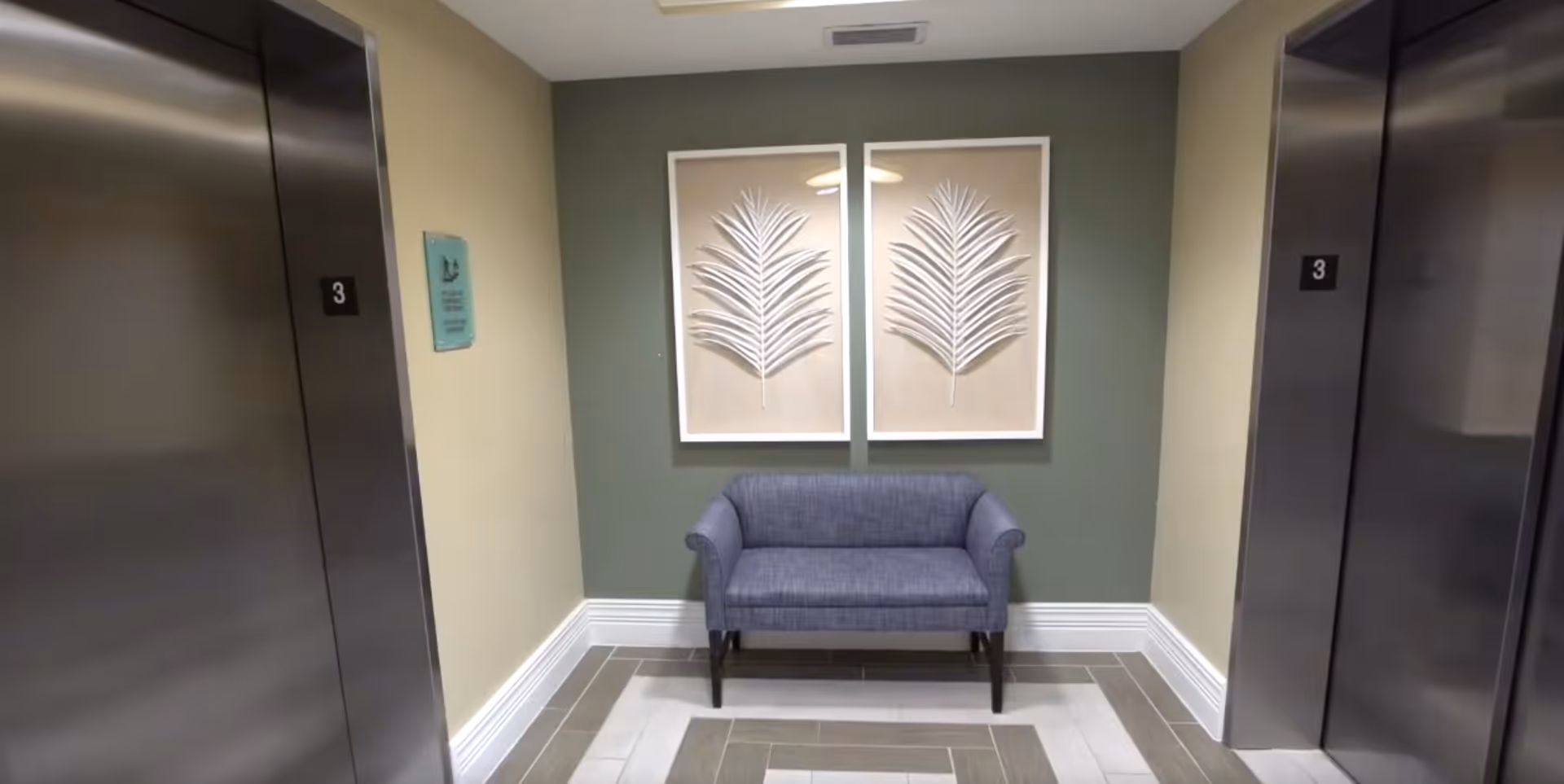A small seating area between two stainless steel elevators with the number 3 on each elevator door. The seating area features a blue upholstered bench against a green accent wall, with two framed leaf artworks hanging above it. The floor has a patterned tile design in shades of gray and white.