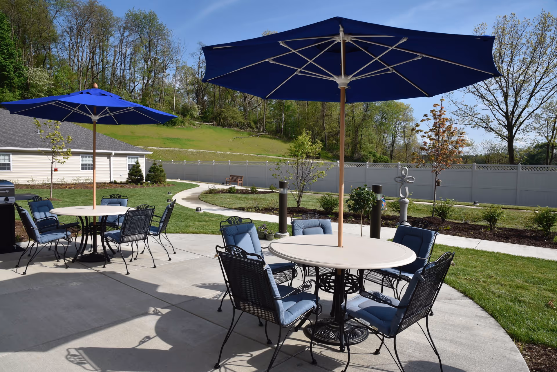 Outdoor patio area with round tables and blue umbrellas, surrounded by cushioned metal chairs. The patio is set on a concrete surface with a grassy lawn and trees in the background under a clear blue sky.