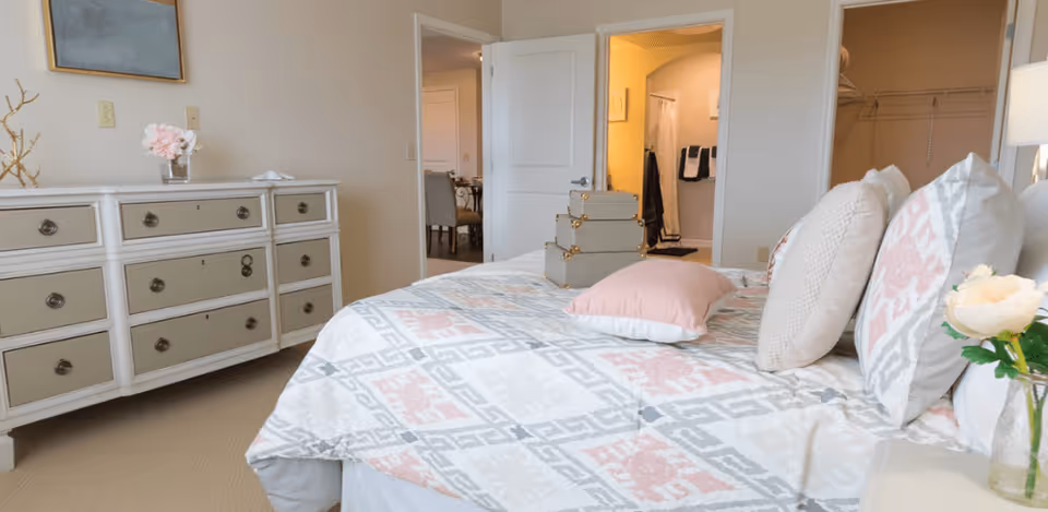 A bedroom with a neatly made bed featuring patterned bedding and several pillows. On the bed are three stacked decorative storage boxes and a pink pillow. To the left is a white dresser with multiple drawers, a vase with pink flowers, and a decorative branch. The room has beige walls and carpet. In the background, an open door reveals a bathroom with towels hanging and a closet with shelving.