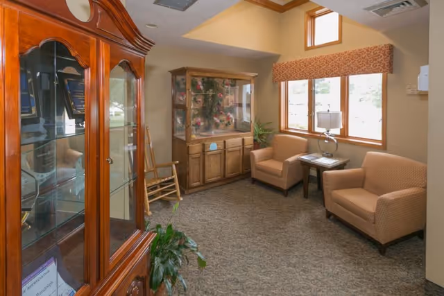 A cozy sitting area in a senior living facility with two cushioned armchairs, a small wooden side table with a lamp, a wooden display cabinet with glass doors containing decorative items, a rocking chair, and a window with a patterned valance letting in natural light.