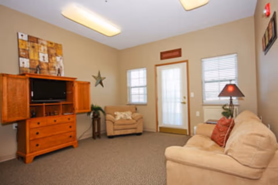 Comfortable living room with beige sofas and armchair, a wooden TV cabinet, lamp, and a door flanked by two windows.