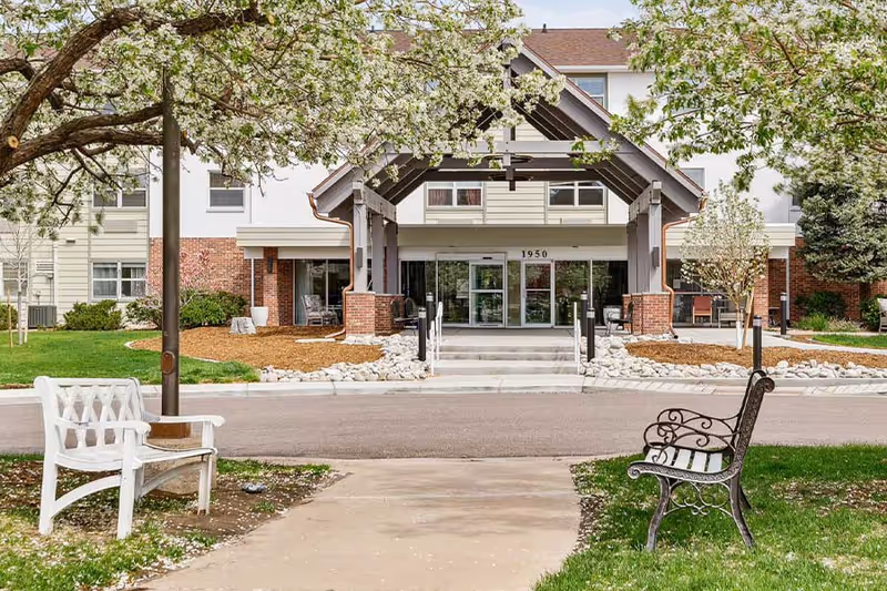 Entrance of a senior living facility with a covered porch supported by wooden beams and brick pillars. There are two benches, one white and one black, on either side of a paved walkway leading to the entrance. Trees with white blossoms frame the scene, and the building has multiple windows and a mix of brick and siding exterior.