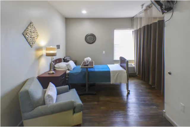 A senior living facility bedroom with a single bed covered with a blue and beige blanket, a bedside table with a lamp, a comfortable armchair with a pillow, a window with blinds, and a wall-mounted TV. The room has wooden flooring and neutral-colored walls with minimal decoration.