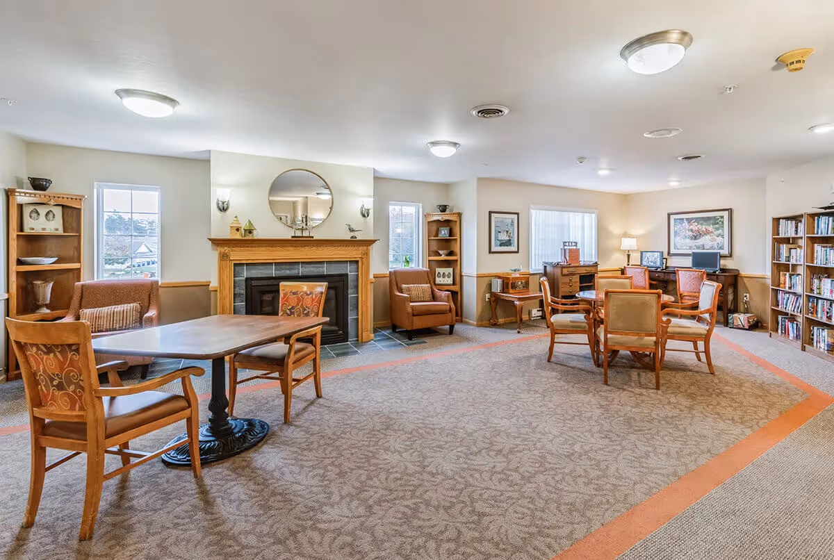 A spacious and well-lit common area in Oak Gardens Place featuring a fireplace with a round mirror above it, several wooden chairs with patterned cushions, tables, bookshelves filled with books, and framed artwork on the walls. The room has carpeted flooring with a decorative border and multiple windows allowing natural light.