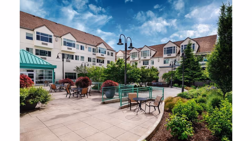 Courtyard patio with tables and chairs, landscaped planters, and a multi-story residential building under a blue sky.
