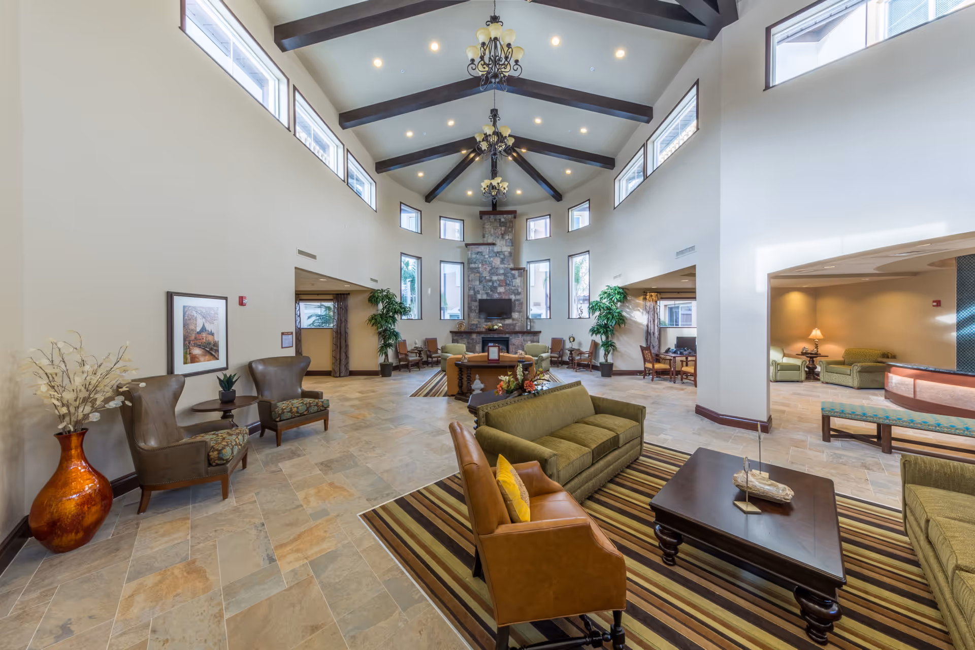 Spacious senior living facility common area with high vaulted ceilings featuring dark wooden beams and multiple chandeliers. The room has large windows near the ceiling and tall windows around a stone fireplace with a mounted TV. The seating area includes green sofas, a brown leather chair with a yellow pillow, striped area rugs, and wooden coffee tables. There are also potted plants, framed artwork on the wall, and additional seating areas with chairs and tables in the background.
