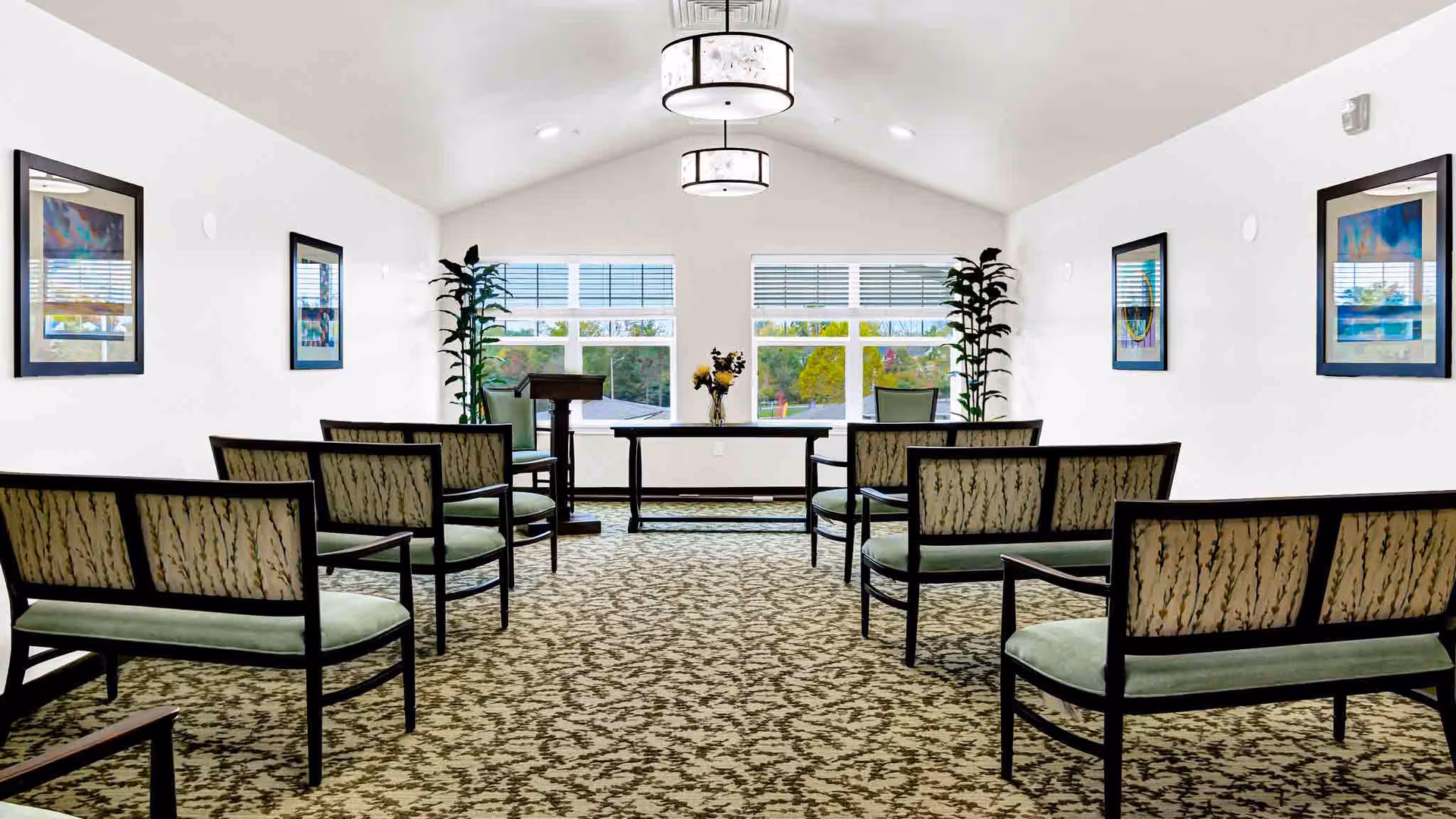 A bright, spacious room with patterned carpet and white walls, featuring rows of green cushioned chairs with dark wooden frames arranged facing a table and podium near large windows with a view of trees outside. Two tall plants flank the windows, and framed artwork hangs on the walls. Two ceiling light fixtures provide illumination.