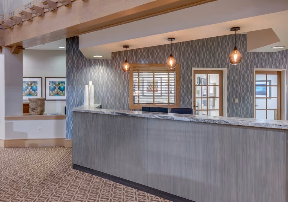 Reception area with a marble countertop desk, three hanging pendant lights, patterned wallpaper, and two doors with glass panels behind the desk. There is a window with blinds behind the desk and framed artwork on the adjacent wall.