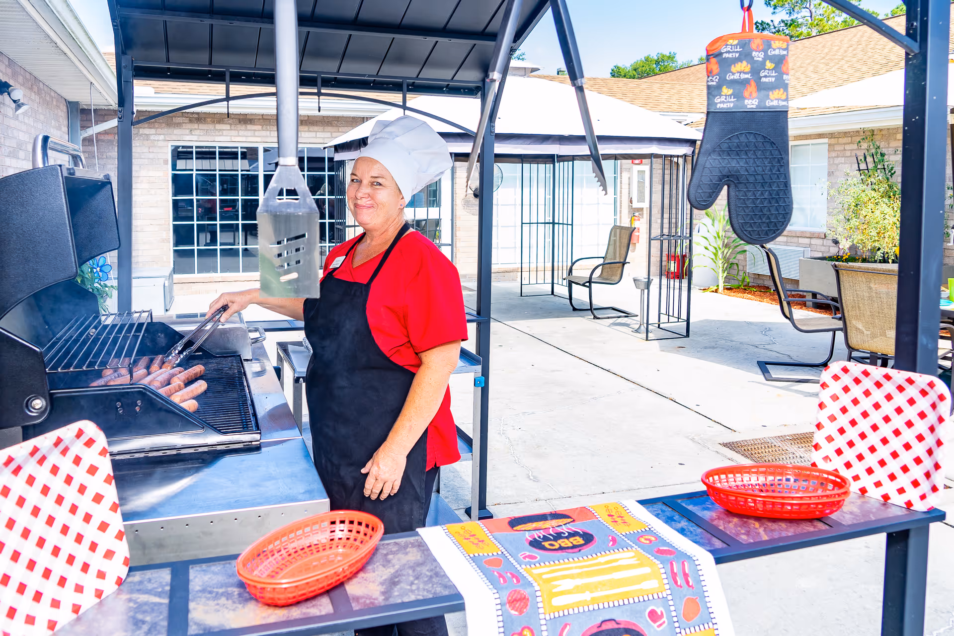 A woman wearing a chef's hat, red shirt, and black apron is grilling sausages on an outdoor barbecue grill under a covered patio area. There are red and white checkered chairs and a table with a colorful BBQ-themed table runner and red baskets nearby. An oven mitt with grill-themed prints hangs from the patio structure. The background shows a building with large windows and patio furniture.