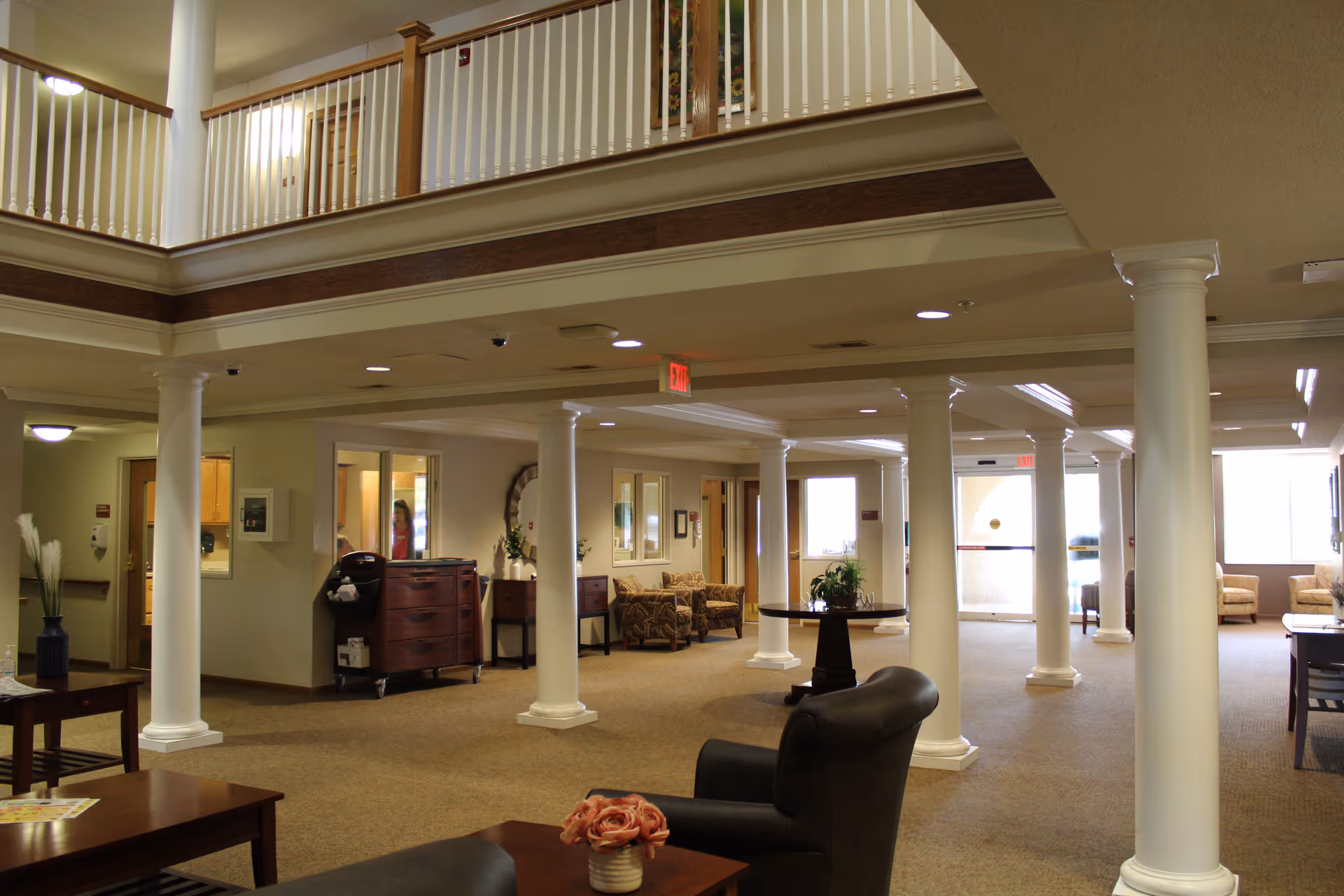 Spacious interior common area of a senior living facility with beige carpet, white columns, and a second-floor balcony railing. The room is furnished with armchairs, tables with flower arrangements, and has multiple doorways and windows letting in natural light.