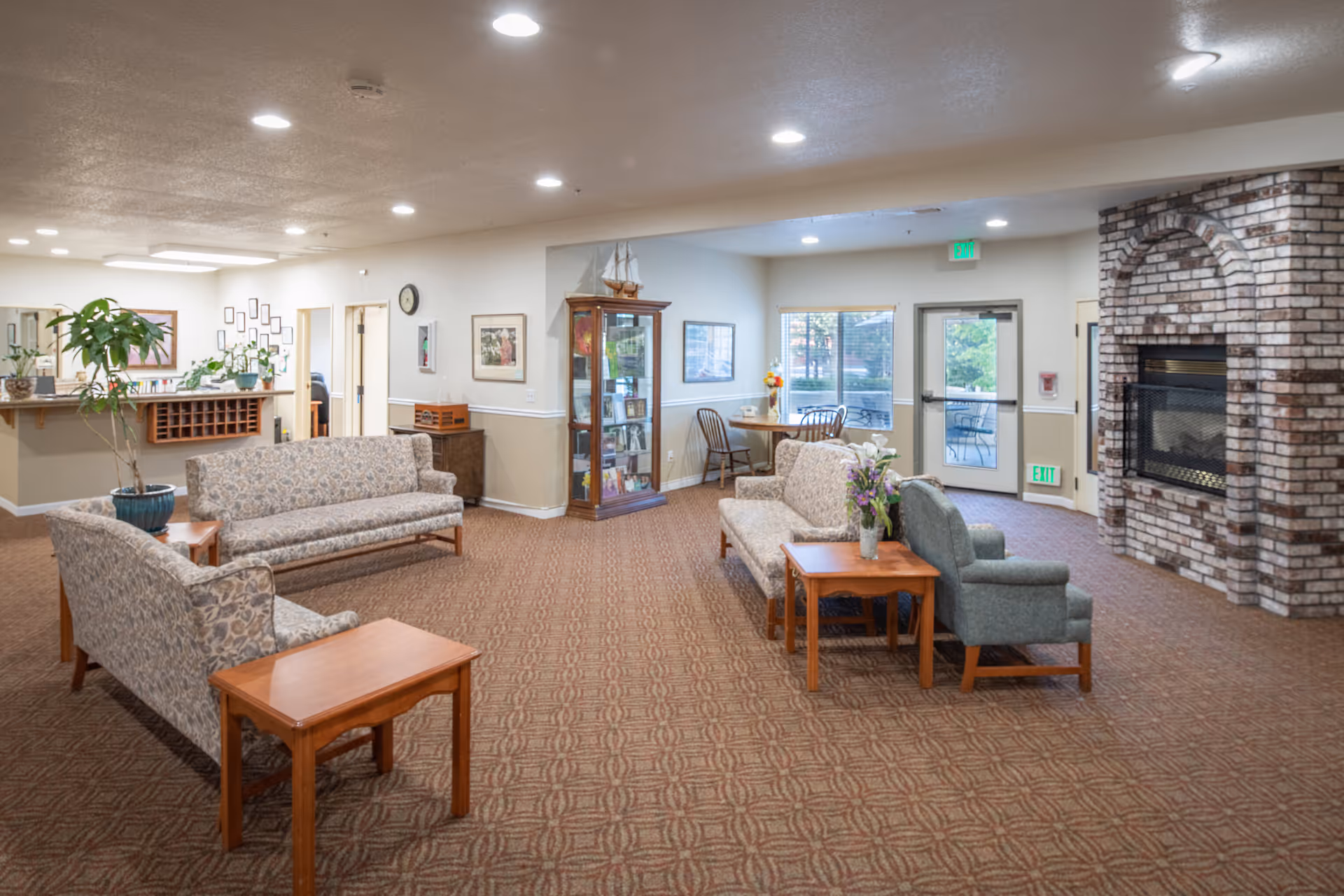 Spacious assisted-living common room with sofas, chairs, side tables, plants, and a brick fireplace.
