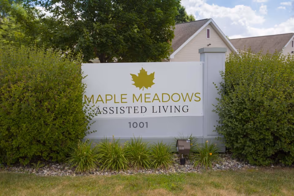 Outdoor sign for Maple Meadows Assisted Living with a maple leaf logo, surrounded by green bushes and grass, with residential buildings in the background under a partly cloudy sky.