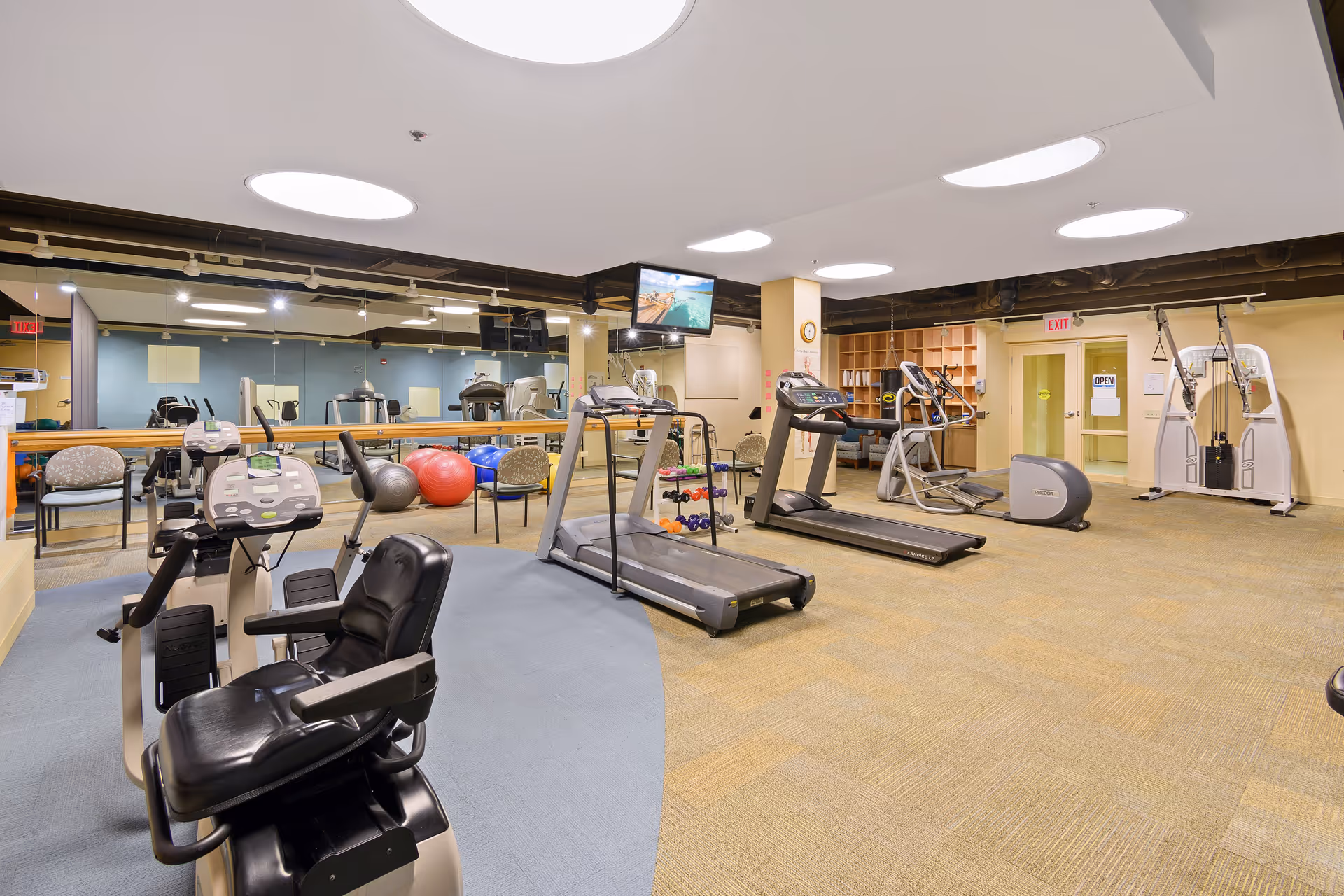 A spacious fitness room in Central Baptist Village featuring various exercise equipment including treadmills, stationary bikes, weight machines, exercise balls, and dumbbells. The room has large mirrors on one wall, circular ceiling lights, and a television mounted on a pillar. There are chairs and shelves along the walls, and the floor is carpeted.