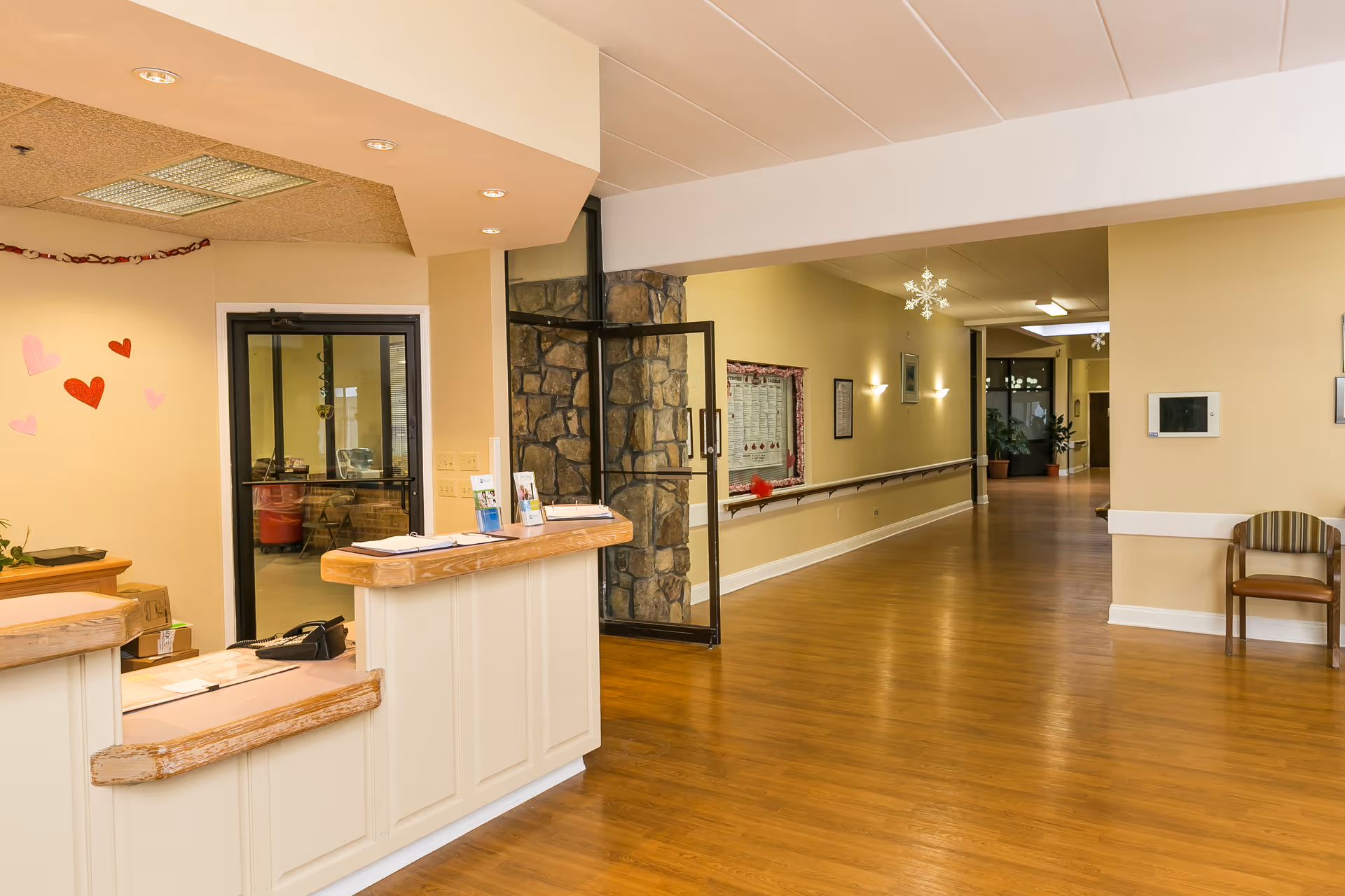 Reception area with a wooden counter and a hallway with wooden flooring and beige walls. The hallway has handrails, wall lights, and decorative snowflakes hanging from the ceiling. There are some heart-shaped decorations on the wall near the reception desk and a chair against the wall on the right side.