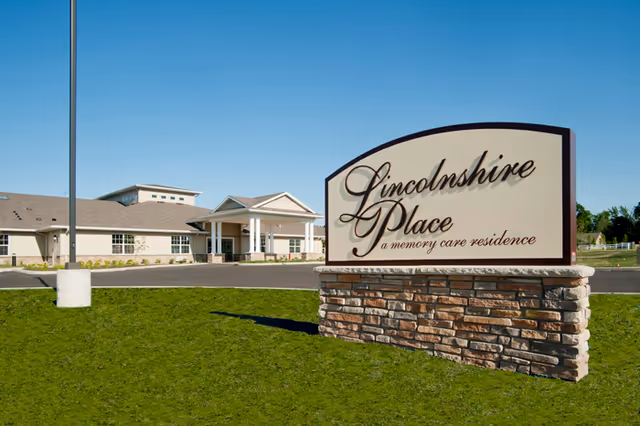 A stone-and-plaque sign reading "Lincolnshire Place a memory care residence" stands on a lawn in front of a single-story assisted living building under a clear blue sky.