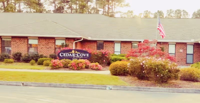 Exterior view of Cedar Cove Assisted Living And Memory Care facility showing a single-story brick building with multiple windows, a manicured lawn, colorful flower beds, bushes, and an American flag on a flagpole.