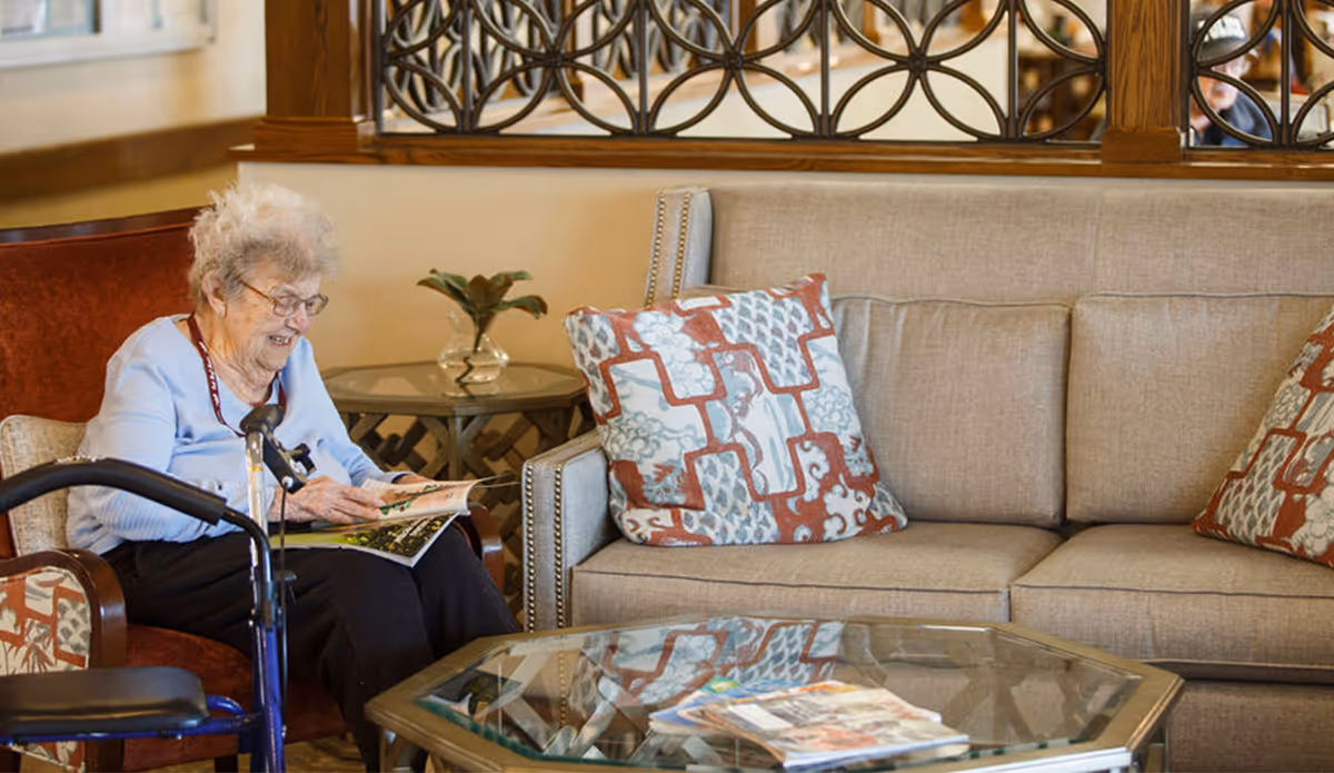 An elderly woman with glasses sitting in a chair next to a beige sofa with patterned cushions, reading a book. There is a glass coffee table with magazines in front of her and a small round side table with a plant behind her. A decorative wooden screen is visible in the background.
