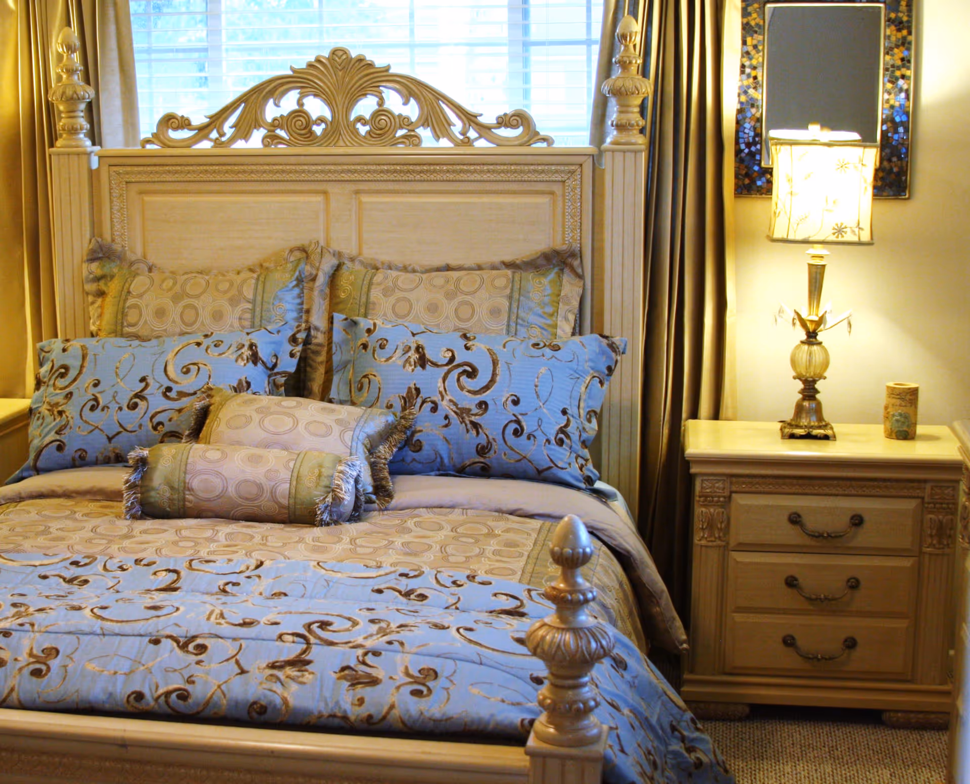 A neatly made bed with an ornate wooden headboard and multiple decorative pillows in blue and beige patterns. Next to the bed is a wooden nightstand with three drawers, a decorative lamp, and a small decorative item. A window with white blinds and beige curtains is behind the bed.