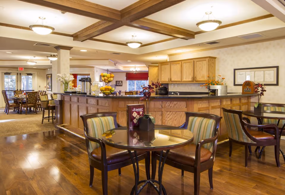 A spacious dining area in a senior living facility with wooden floors, round glass-top tables with striped cushioned chairs, and a wooden counter with cabinets. The room is well-lit with ceiling lights and has decorative plants and a fruit stand on the counter. In the background, there are more dining tables and large windows letting in natural light.