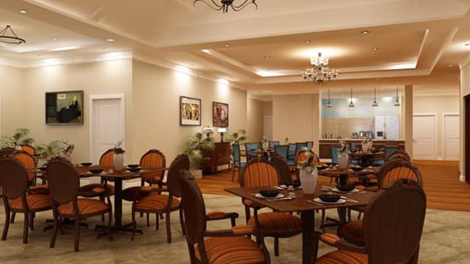 A spacious dining room with multiple wooden tables and chairs featuring orange striped upholstery. The room is warmly lit with ceiling lights and chandeliers. There are framed pictures on the walls and some potted plants placed around the room. In the background, there is a kitchen area with pendant lights hanging above a counter.