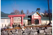 Exterior view of Kin On Healthcare Center building with a stone wall and landscaped plants in front. The building has a green roof and a covered entrance with red pillars.