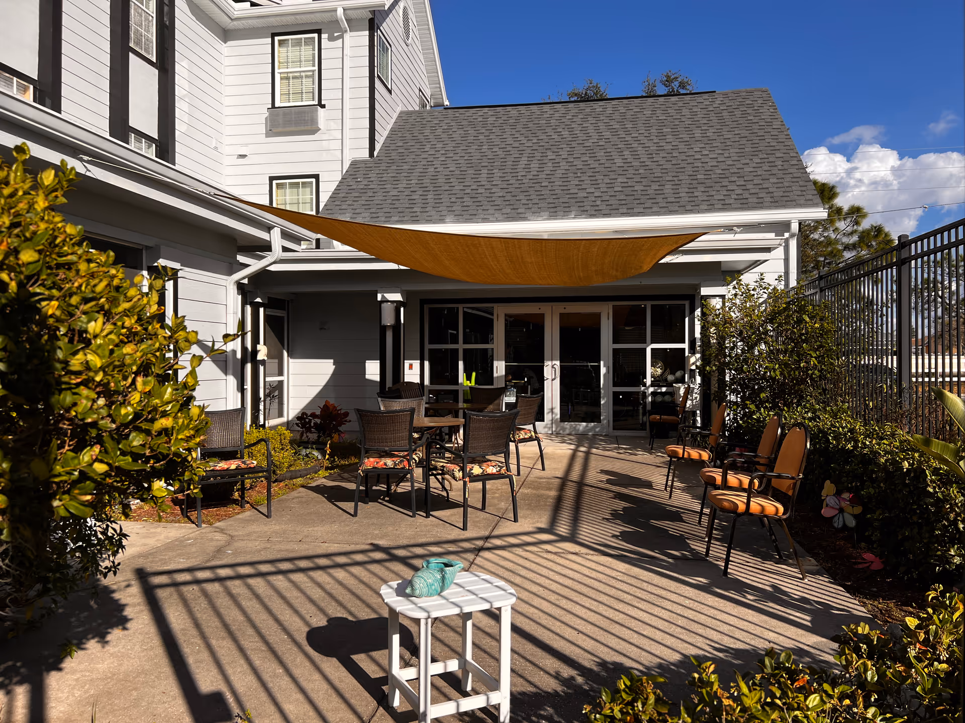 Sunlit outdoor patio courtyard with tables, chairs, a triangular shade sail and glass doors leading into the white building.