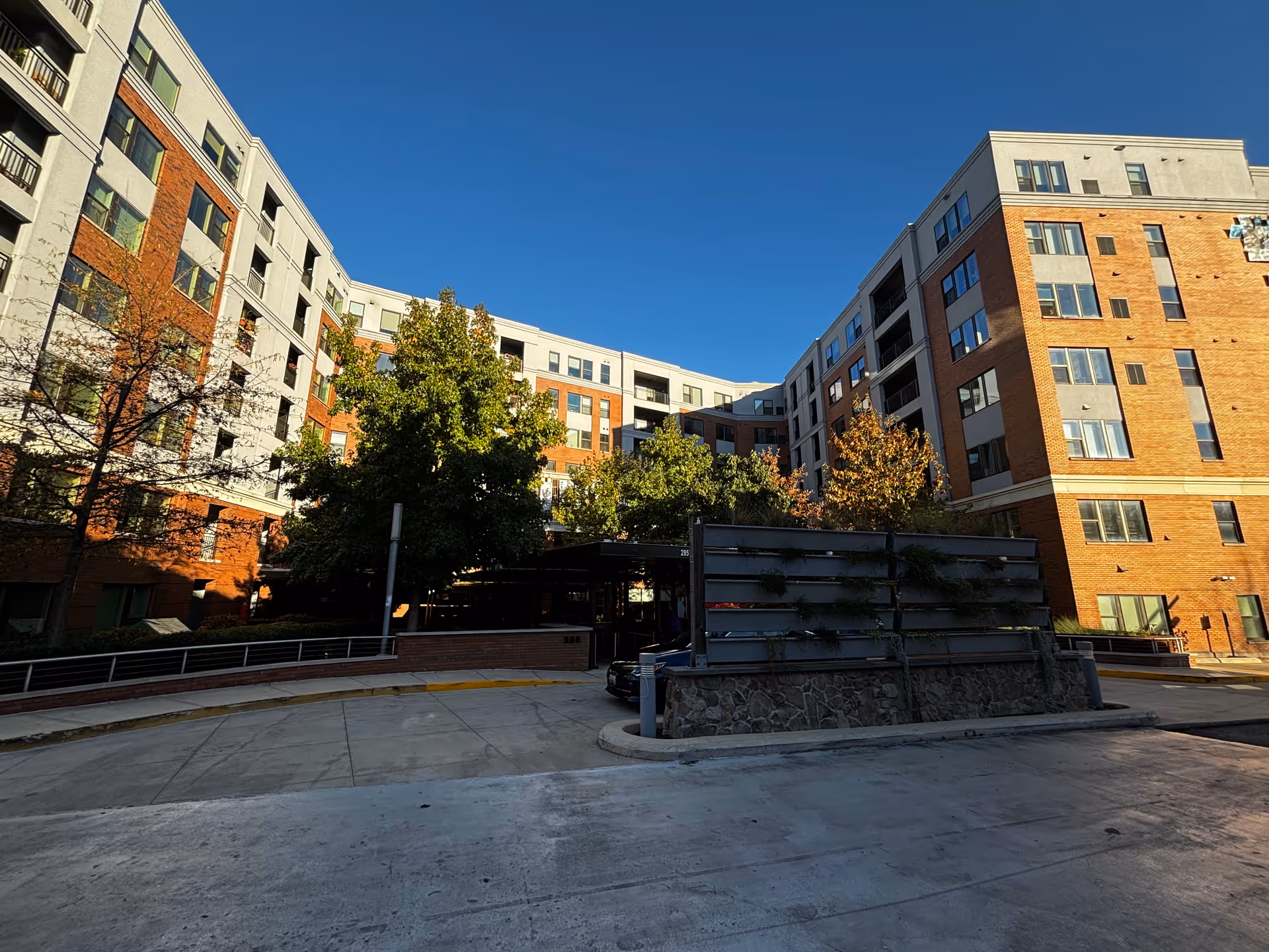 Exterior view of a multi-story senior living facility building with a mix of brick and light-colored walls, surrounded by trees and a clear blue sky.