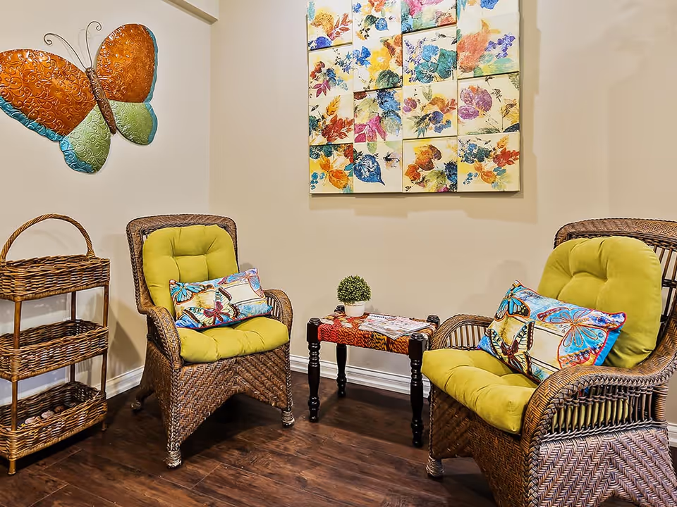 A cozy sitting area with two wicker chairs featuring green cushions and butterfly-themed pillows. Between the chairs is a small table with a colorful cloth, a small potted plant, and some magazines. On the wall behind the chairs is a large colorful artwork composed of multiple panels with floral and leaf designs. To the left, there is a wicker shelf and a large decorative butterfly wall art.