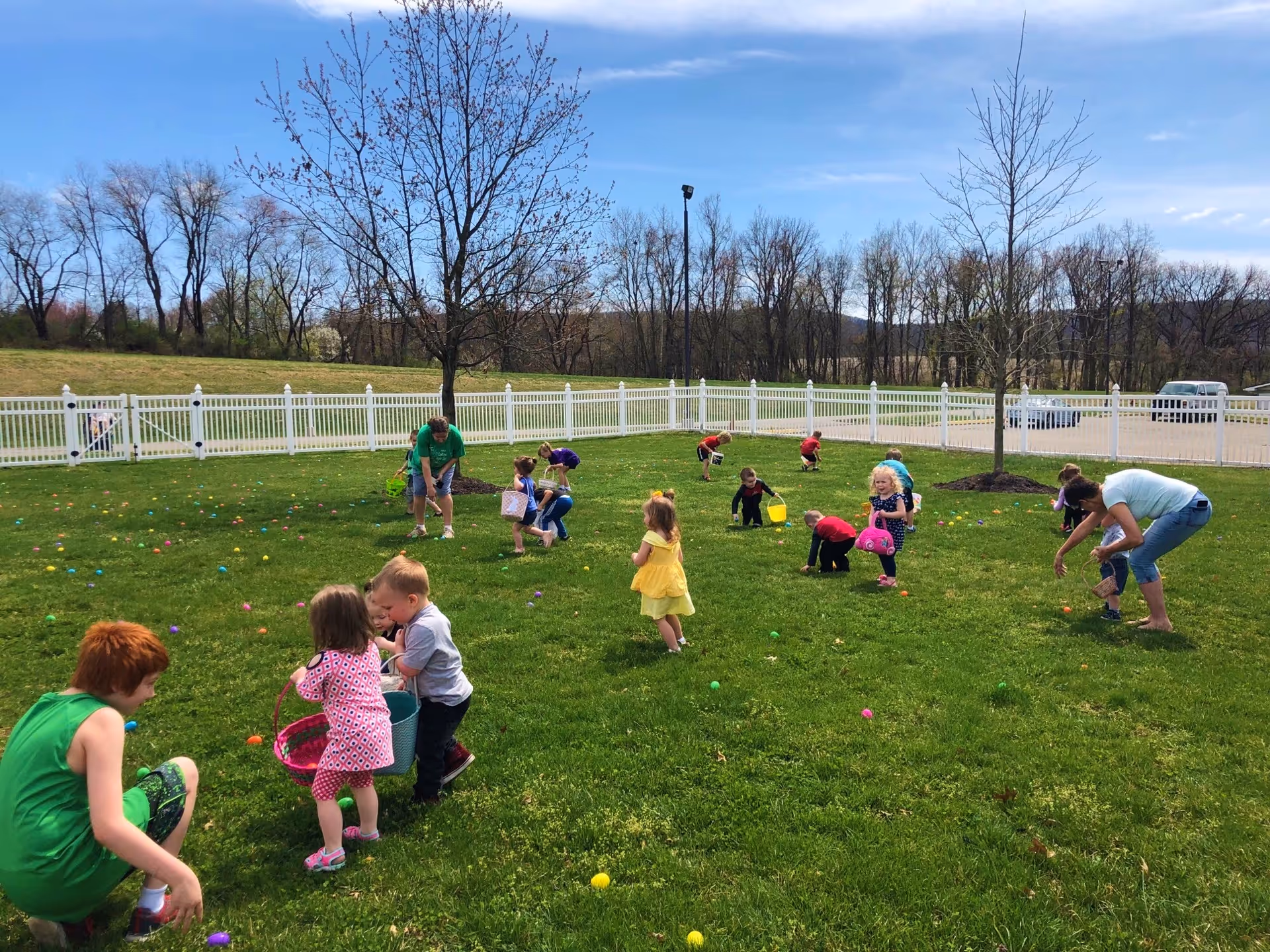 Children and a few adults participating in an Easter egg hunt on a grassy lawn enclosed by a white picket fence, with leafless trees and a clear blue sky in the background.