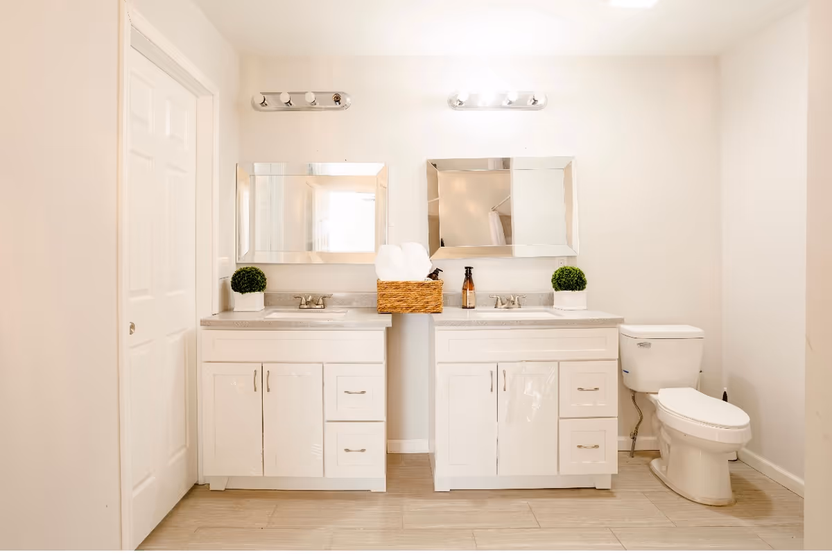 A bright bathroom featuring two white vanities with sinks, each topped with a rectangular mirror and a light fixture above. A wicker basket with rolled white towels sits between the vanities. There are small green potted plants on each vanity. To the right, there is a white toilet. The floor is light-colored tile, and the walls are painted white.