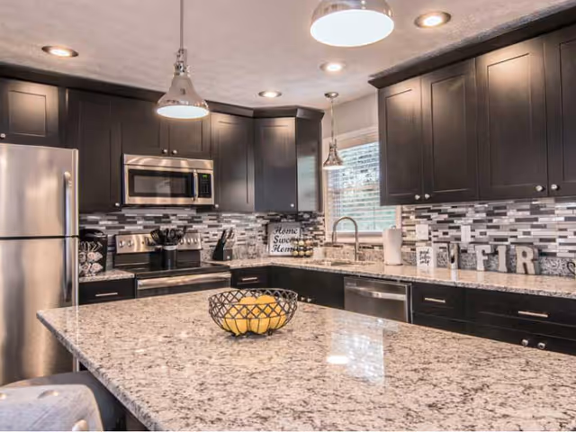 Modern kitchen with dark wood cabinets, stainless steel refrigerator, microwave, and stove. Granite countertops with a large island in the center holding a wire fruit basket with lemons. A window above the sink lets in natural light, and the backsplash features a mosaic tile design in shades of gray and white.