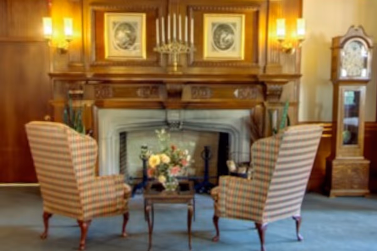 A cozy sitting area with two plaid upholstered armchairs facing a wooden fireplace mantel. The mantel is decorated with a candelabra and two framed pictures. A small wooden table with a floral arrangement is placed between the chairs. To the right of the fireplace stands a tall grandfather clock. The room has warm wooden paneling and soft lighting from wall sconces.