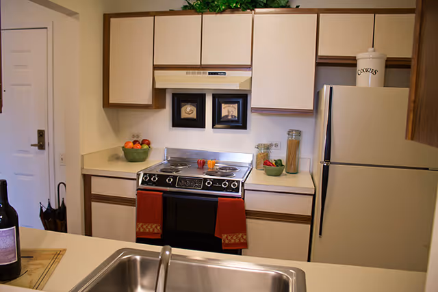 A compact kitchen with beige cabinets and countertops, featuring a stove with two red towels hanging from the handle, a refrigerator with a cookie jar on top, a sink in the foreground, and decorative items including framed pictures, a bowl of fruit, and jars of pasta and vegetables on the counter.