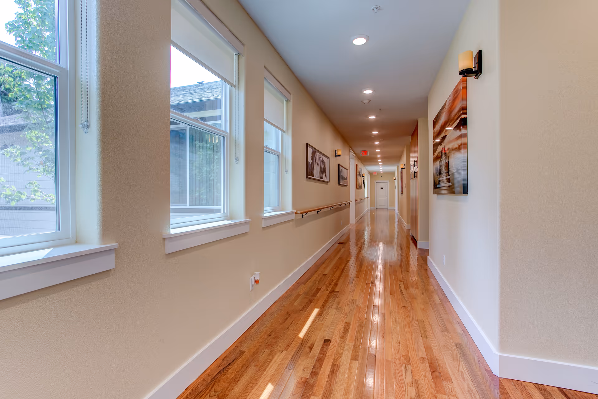 A long, well-lit hallway with wooden flooring and beige walls. On the left side, there are several large windows with white frames and roller blinds, allowing natural light to enter. On the right side, there are framed pictures and wall-mounted light fixtures. A handrail runs along the left wall beneath the windows. The hallway extends to a white door at the far end.